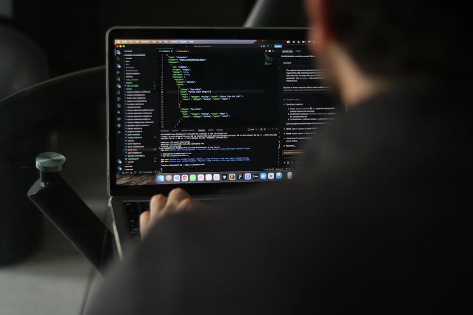 A person using a laptop, viewed from behind, displaying lines of code on the screen in a dark environment. The individual is seated, with their right hand on the keyboard, and the laptop shows a dark-