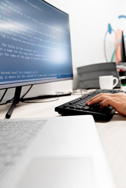 A close-up view of a person's hands working on a laptop computer placed on a dark tabletop. The screen displays lines of colorful code with a black background, indicating programming or web developmen